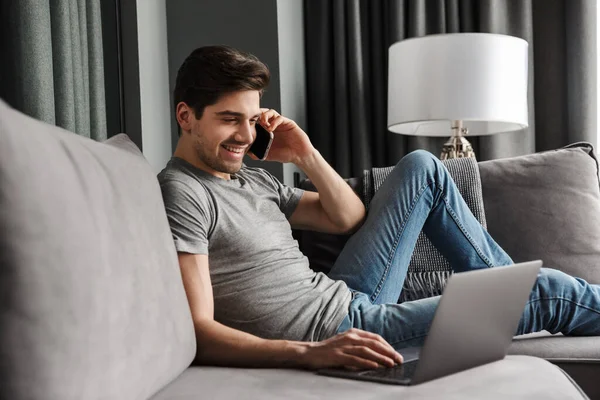 リビングルームのソファに腰を下ろし ノートパソコンを使いながら携帯電話で話す魅力的な笑顔の若い髭の男の肖像画 — ストック写真