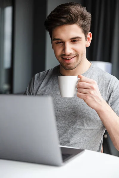 家のテーブルに座ってコーヒーを飲みながらラップトップコンピュータを使ってカジュアルな服を着ているハンサムな笑顔の若い髭の男 — ストック写真