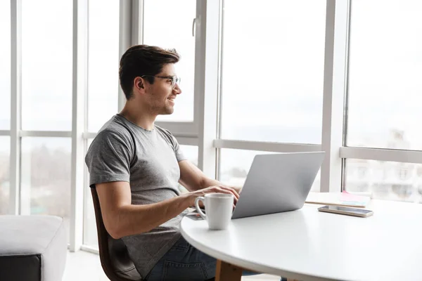 年轻英俊的留着胡子的男人坐在桌旁 穿着休闲装 一边用笔记本电脑 一边笑 — 图库照片