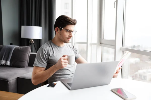 年轻英俊的留着胡子的男人坐在桌边喝咖啡时 穿着休闲装 用笔记本电脑 — 图库照片