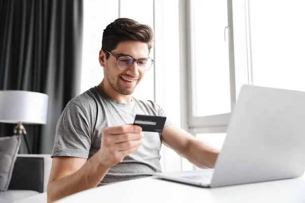 stock image Handsome smiling young bearded man wearing casual clothes using laptop computer while sitting at the table at home, showing plastic credit card, shopping