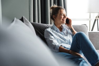 Çizgili tişörtlü mutlu genç bir kadının oturma odasında kanepede otururken gülüp cep telefonu kullanması.