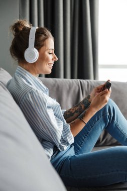 Oturma odasındaki kanepede otururken cep telefonu ve kulaklık kullanan çizgili tişörtlü gülümseyen genç bir kadın.