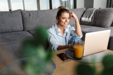 Oturma odasında dizüstü bilgisayar kullanan, kulaklıklı, gülümseyen bir kadın görüntüsü.