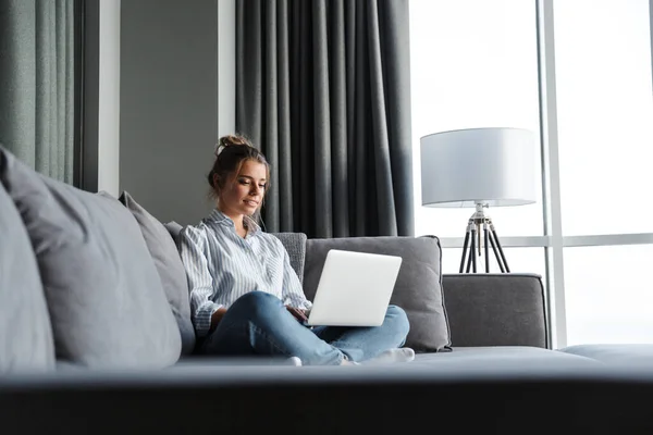 Image of pleased nice woman smiling and using laptop while sitting on couch at living room