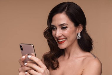 Bej arka planda izole edilmiş, gülümseyen ve cep telefonu tutan muhteşem, yarı çıplak bir kadının güzel portresi.