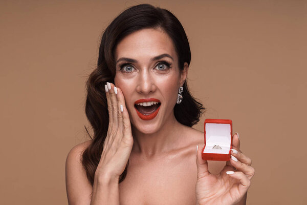Красота портрет счастливой великолепной взрослой полуобнаженной женщины с коробкой обручального кольца изолированы на бежевом фоне
