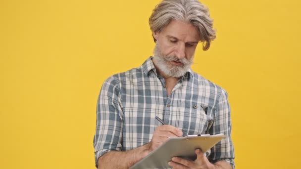 Senior Concentrado Homem Isolado Sobre Fundo Amarelo Segurando Prancheta — Vídeo de Stock