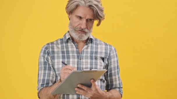 Senior Ernstige Man Geïsoleerd Gele Achtergrond Met Klembord Schrijven Notities — Stockvideo