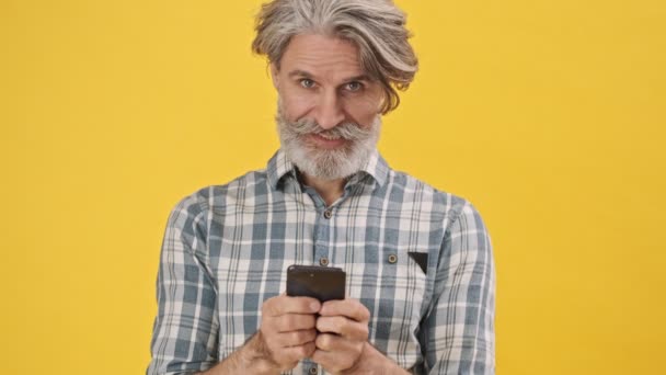 陽気な正シニア男隔離上の黄色の背景使用して携帯電話チャット — ストック動画