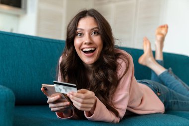 Evde koltukta cep telefonu kullanan, şaşırmış, genç bir kadının resmi..