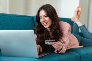 Evde kanepe üzerinde kredi kartı tutan dizüstü bilgisayar kullanan pozitif duygusal genç bir kadının görüntüsü.