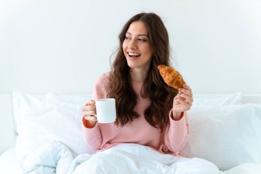 Evde, yatak odasında, yatakta, kruvasan ve kahveyle iyimser genç bir kadının görüntüsü..