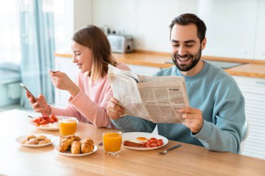 Evde rahat bir mutfakta kahvaltı ederken cep telefonu kullanan ve gazete okuyan genç çiftin portresi.