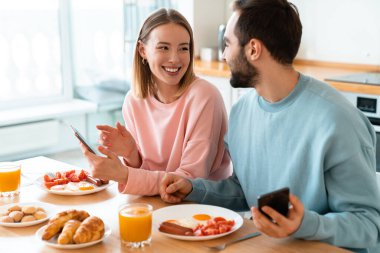 Evde rahat bir mutfakta kahvaltı ederken cep telefonu kullanan ve gülen genç çiftin portresi.