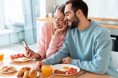 Evde rahat bir mutfakta kahvaltı ederken cep telefonu kullanan ve gülen genç çiftin portresi.
