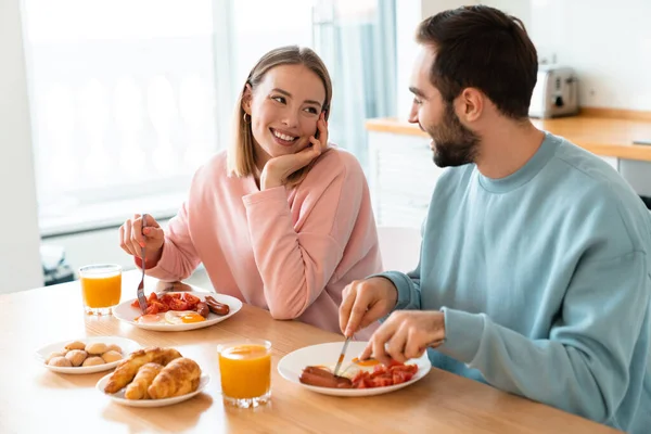 若い幸せなカップルの肖像画自宅で居心地の良いキッチンで朝食を食べながらテーブルで一緒に食べる — ストック写真