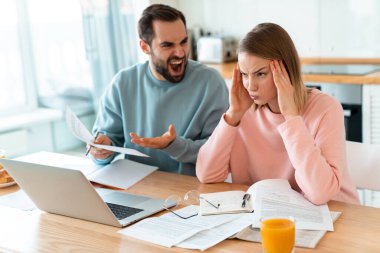 Evde dizüstü bilgisayarla ve belgelerle çalışırken tartışan genç çiftlerin portresi.