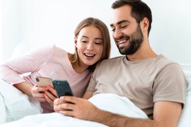 Evdeki yatak odasında yatarken gülümseyen ve akıllı telefon kullanan güzel çiftin portresi.