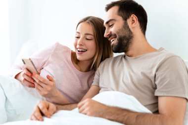 Evdeki yatak odasında uzanırken gülümseyen ve akıllı telefon kullanan güzel çiftin portresi.