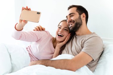 Güzel, neşeli bir çiftin portresi. Evde, yatak odasında uzanmış, akıllı telefonlarda selfie çekiyorlar.