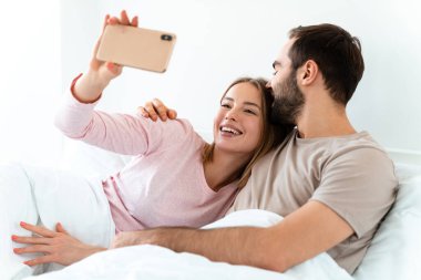 Güzel, neşeli bir çiftin portresi. Evde, yatak odasında uzanmış, akıllı telefonlarda selfie çekiyorlar.