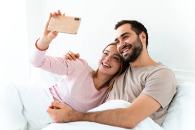 Güzel, neşeli bir çiftin portresi. Evde, yatak odasında uzanmış, akıllı telefonlarda selfie çekiyorlar.