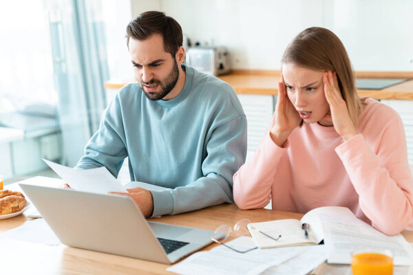 Портрет молодой задумчивой пары, работающей с ноутбуком и документами на уютной кухне дома
