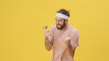 Stüdyoda, sarı bir arka planda izole edilmiş sevinçten havalara uçarken, spor kıyafetleri içinde, mutlu, genç bir sporcu elinde bir ödül tutuyor.