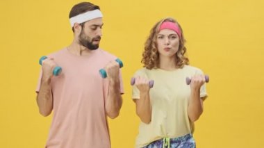 Spor kıyafetleri içinde genç bir çift spor yapıyor. Stüdyonun sarı arka planında izole haldeki dambılları kaldırıyor.