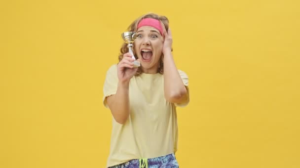 スタジオで黄色の背景に隔絶された賞を持ちながら スポーツウェアに身を包んだ幸せな若いアスリート女性が喜んでいる — ストック動画