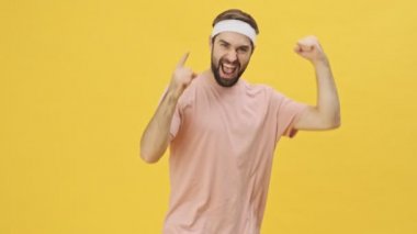 Spor kıyafetleri içinde mutlu, genç bir atletik adam kamerayı işaret ediyor ve kollarını kaldırırken stüdyodaki sarı arka planda izole bir şekilde kaslarını gösteriyor.