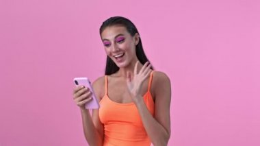 Pembe duvarın üzerinden izole edilmiş genç ve mutlu bir kadın cep telefonuyla görüntülü konuşma yapıyor.