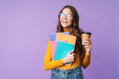 Gözlük takan, elinde egzersiz kitapları ve menekşe arkaplanından izole edilmiş kahve fincanı tutan güzel bir öğrenci resmi.