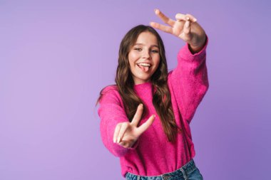 Uzun kahverengi saçlı, güzel bir kadının görüntüsü gülümseyen ve barış işareti yapan parmaklar menekşe arka planda izole edilmiş.