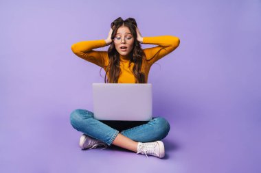 Dizüstü bilgisayar kullanan genç ve güzel bir kadının görüntüsü. Bacakları çapraz otururken menekşe arkaplan üzerinde izole edilmiş.