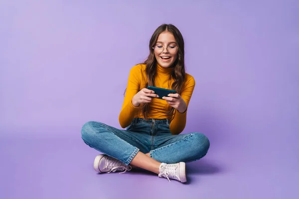 足で座っている間にスマートフォンを持っている若い美しい女性のイメージは 紫色の背景に隔離された交差 — ストック写真