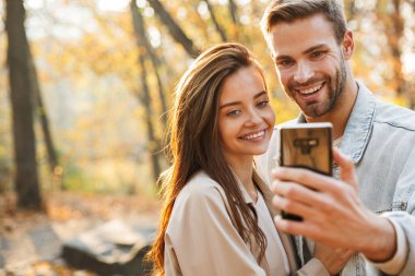Genç ve güzel bir çift, sonbahar parkında selfie çekerken cep telefonu kullanıyor.