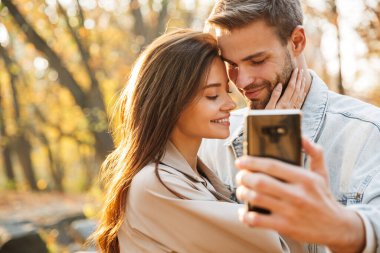 Genç ve güzel bir çift, sonbahar parkında selfie çekerken cep telefonu kullanıyor.