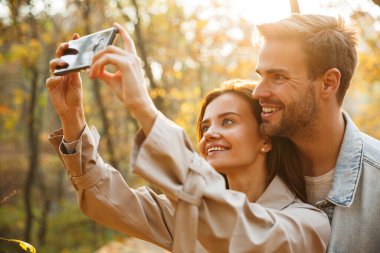 Genç ve güzel bir çift, sonbahar parkında selfie çekerken cep telefonu kullanıyor.