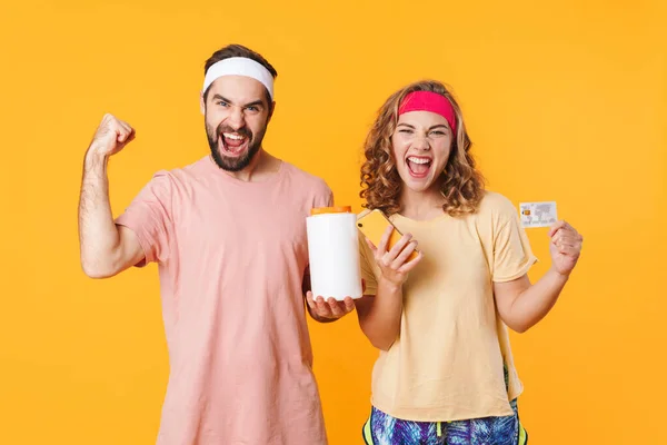 Portrait of young happy caucasian fitness couple wearing headbands holding protein jar and credit card isolated over yellow background