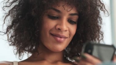 Genç pozitif Afrikalı kıvırcık kadın evde cep telefonu kullanıyor.