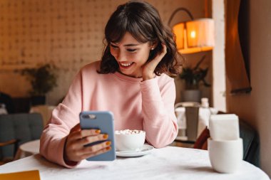 Kahvede cep telefonuyla kahve içen mutlu, esmer bir kadının fotoğrafı..