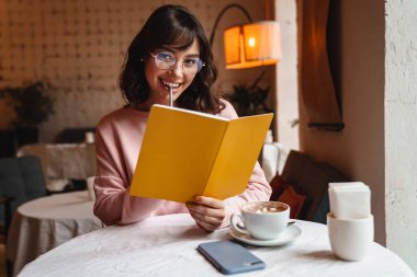 Güzel, mutlu, gülümseyen bir esmer kadının fotoğrafı. Kapalı bir kafede kitap okuyan genç bir kadın..