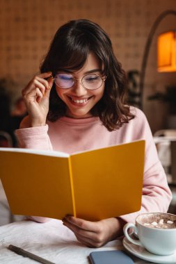 Kafede kitap okuyan güzel, pozitif gülümseyen, esmer bir kadının resmi..