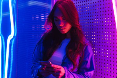 Neon bir duvarın önünde cep telefonu kullanarak duran uzun siyah saçlı güzel Asyalı kadın.