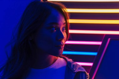 Uzun siyah saçlı, güzel Asyalı bir kadın neon bir duvarın önünde dikiliyor, başka tarafa bakıyor.