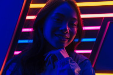 Güzel, gülümseyen, uzun siyah saçlı Asyalı bir kadın neon bir duvarın önünde poz veriyor.