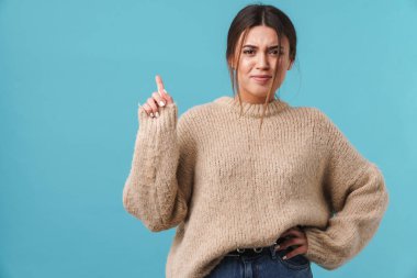 Süveter giymiş kafası karışmış genç bir kadının mavi arka planda parmakla işaret etmesi.