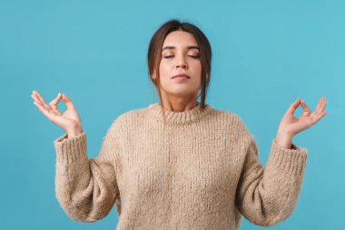 Sakin, meditasyon yapan ve parmaklarını bir arada tutan güzel bir kadının görüntüsü mavi arka planda izole edilmiş.